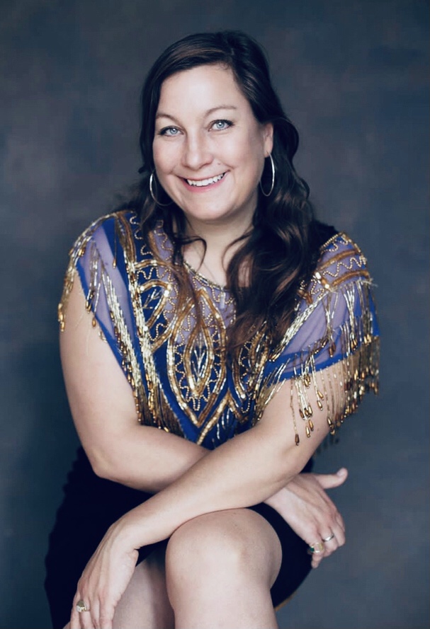 A photo of Caroline McGlone who has dark brown long hair, she has pale skin and is wearing a blue short sleeve blouse with gold embellishment and a black skirt. She is smiling to camera. 